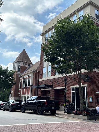 The image depicts a street scene in Naperville, Illinois, United States of America. A black GMC Sierra pickup truck is parked on the right side of the street, facing away from the camera. To its left, a black SUV is also parked. Further down the street, more cars are visible. The buildings lining the street are made of brick, with some featuring large windows and architectural details like a conical tower. A large tree with dense green foliage stands prominently in the foreground, partially obscuring the view of the building behind it. A woman is seated on a bench outside one of the buildings. The sky is partially cloudy.