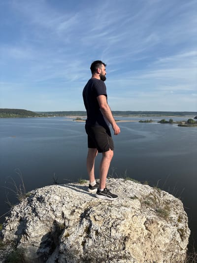 The image depicts a man standing on the edge of a rocky cliff overlooking a large body of water. He is wearing a dark t-shirt and shorts, and athletic shoes. He is looking upwards. The sky is blue with wispy clouds. The body of water appears to be a river or lake, with land visible in the distance. The location is in the vicinity of Gorbunyata, Russia, as deduced from the geographical features visible in the background.