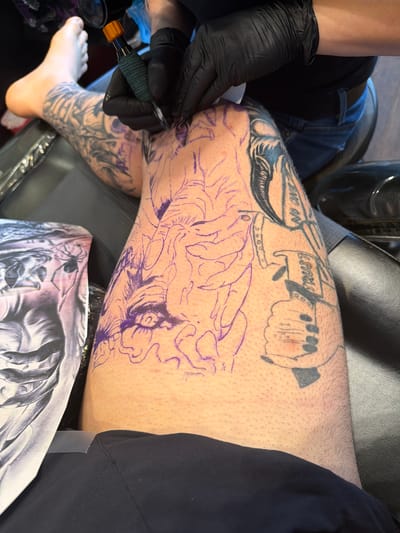 A person is getting a tattoo on their leg, likely in Arlington Heights, Illinois, USA. The tattoo artist, wearing black gloves, is using a tattoo machine on the person's right thigh. A stencil of the tattoo design is visible, and there are pre-existing tattoos above the new design. A reference picture for the tattoo is visible on a surface near the leg. The person is sitting on a chair with what looks like a black outfit.