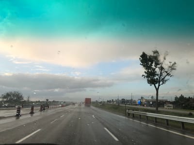 The image depicts a view from inside a vehicle traveling on a highway in Innisfil, Canada. The scene is set on a wet, overcast day, likely during the late afternoon or early evening, indicated by the soft, diffused light and the colors of the sky. The road is wet, with visible reflections of traffic and the sky, suggesting recent or ongoing rain.

The highway appears to be undergoing construction, with orange and black traffic barrels lining the left side of the road. Several vehicles are visible on the highway, including cars and a large orange truck in the distance. A lone tree with sparse foliage stands on the right side of the road, silhouetted against the cloudy sky. Beyond the highway on the right, there are commercial buildings with visible signage. One sign reads "LEGEND" in large letters, and another smaller sign appears to indicate a speed limit of "100". Other signs are partially visible, suggesting a commercial area or rest stop. The sky is a mix of grays and blues, with a noticeable teal hue dominating the upper portion of the image, possibly due to atmospheric conditions or a filter. The overall mood is somewhat somber due to the weather, but the ongoing activity on the highway suggests normal traffic flow. Water droplets are visible on the windshield, reinforcing the wet weather conditions.