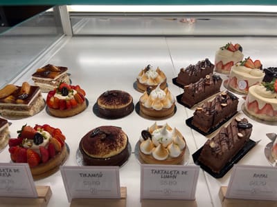 The image shows a glass display case of pastry desserts in a bakery, located at 9.063 latitude, -79.391 longitude in Panama City, Panama. The pastry display case contains various types of desserts, including tarts, cakes, and pastries. The case is lit from above, highlighting the various textures and colors of the desserts. The bakery is a high-end establishment, with a modern and elegant design. The desserts are arranged in a visually appealing manner, with a variety of colors and textures. The pastry display case is empty on the left, but the right side is filled with a variety of pastry desserts. Each dessert has a small sign with the name and price. The desserts are arranged in rows, with the larger cakes on the bottom and the smaller desserts on the top. There are a total of 12 pastries in the display case. The pastries are all freshly baked, with a variety of flavors and textures. The bakery appears to be clean and well-maintained. There are no people in the image.