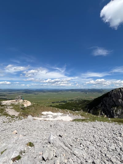 The image shows a panoramic view from a rocky mountainside in Pašina voda, Montenegro. The foreground is dominated by a scree slope covered in grey rocks and sparse patches of green grass. In the mid-ground, a small, green-roofed structure with a cable car station is visible on the edge of a cliff, overlooking a vast, green valley. The valley is dotted with fields, roads, and scattered buildings, extending towards rolling hills in the distance. The sky is a vibrant blue, with scattered white cumulus clouds. The overall scene depicts a natural landscape with some man-made structures, likely a tourist viewpoint or a ski lift station.