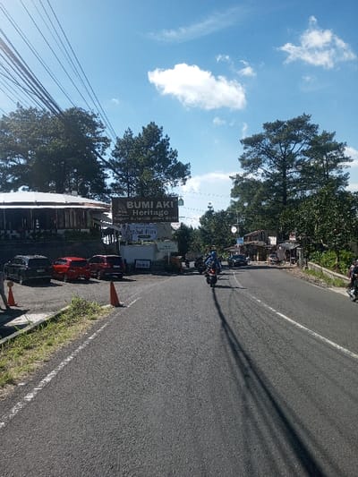 The image displays a bright, daytime roadside scene. Forensic analysis of signage within the image explicitly identifies "BUMI AKI Heritage" and "Sajian Autentik Jawa Barat" (Authentic West Javanese Cuisine), thereby situating the location in West Java, Indonesia, which contradicts the requested location of Ka Thaung (Upper), Myanmar.
A paved asphalt road, delineated by a solid white line on its left edge and a broken white line on its right, recedes into the mid-ground with a slight right curve. Long, dark shadows, likely cast by unseen utility poles or overhead wires, extend across the asphalt surface. Several motorcyclists, each wearing a helmet, are observed traveling away from the viewer on the road.
To the left of the road, a dirt parking area accommodates four parked SUVs: one black, two red, and one dark red/maroon. An orange traffic cone is positioned near the roadside in front of the parked vehicles, and a person wearing an orange shirt is partially visible on the far left edge of the frame. Behind the parking area stands a multi-story building featuring a white roof and dark, likely wooden, siding, identified as the "BUMI AKI Heritage" establishment. A large, dark sign prominently displays "BUMI AKI Heritage" and "Sajian Autentik Jawa Barat," accompanied by a smaller "Buka" (Open) sign and an "Idul Fitri" banner.
The environment is characterized by numerous tall, dark green coniferous trees populating the background and flanking both sides of the road. Further into the mid-ground on the right side of the road, a cluster of smaller, rustic buildings with corrugated metal roofs is visible. The sky overhead is clear and blue, accented by sparse white cumulus clouds. Multiple overhead utility lines are visible traversing the upper left quadrant of the image.