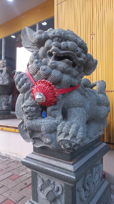 A grey stone Chinese guardian lion (Foo Dog) statue is positioned outdoors on a multi-tiered grey stone pedestal, which features carved decorative elements including a lattice pattern. The statue displays intricate carvings depicting its mane, teeth, and a smaller lion figure at its base. A red ribbon with a prominent red, circular, pleated ornament centered with a silver element is tied around the lion's neck. The ground surface consists of red and grey interlocking pavers. In the background, a building with a closed, bright yellow corrugated or slatted rolling door is visible, alongside reflective window panes through which bright interior lights are apparent. A portion of another identical grey stone guardian lion statue is visible to the left, indicating a pair. The scene is consistent with the entrance of a Chinese temple or a building within a Chinese cultural district, specifically located in Glodok, West Jakarta, Special Capital Region of Jakarta, Indonesia. No people, active subjects, or interactions are present.