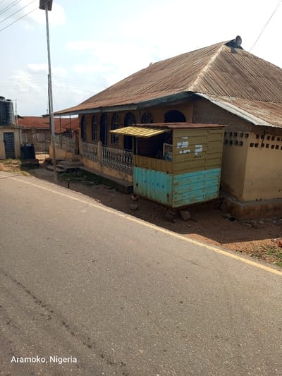 The image shows a street in Aramoko, Nigeria, located at latitude 7.84 and longitude 4.485. There is a single-story building with a corrugated metal roof on the right side of the street. The building has a partially covered awning and an open storefront with a small, rusted, blue and yellow metal shed in front. The building is located on the right side of the street and is surrounded by a dirt area and grass. The street has a yellow dividing line in the center, and a black power pole extends vertically on the left side of the street. There is a blue water tank on the left side of the street. The pavement on the street appears to be cracked and worn. There is a small amount of debris visible on the ground. There are no people or vehicles visible in the image. The building is likely a small shop or store. The shed in front of the building could be a storage space for merchandise or tools. The power pole suggests that there is electricity available in the area. The blue water tank may be used for storage or for providing water for the neighborhood. The overall scene suggests that the image was taken in a rural or suburban area.