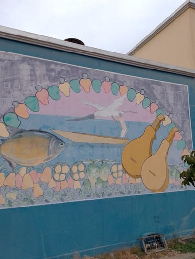 A mural is painted on a teal-colored wall. The mural depicts a coastal scene with a large fish on the left, a person in a boat with a bird flying overhead in the center, and two stringed instruments on the right. The background of the mural features a gray, urban landscape with buildings and a curved border of colorful fruit or vegetable shapes. The sky in the mural is a soft pink and blue. In front of the teal wall, a metal grate covers a drainage opening on the ground. The wall itself has some graffiti or markings near the bottom right. The mural is located on the side of a building, with part of a yellow building visible above the mural on the right side. The sky above is overcast.