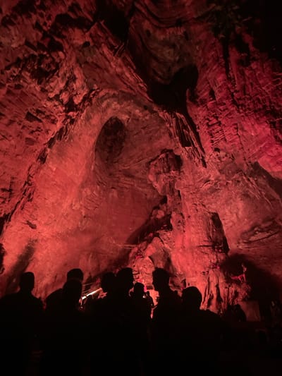 The image depicts the interior of a dimly lit cave, illuminated by a vibrant red light. The cave walls are rough and textured, displaying distinct layers and formations typical of a natural cavern. In the foreground, silhouetted figures of people are visible, suggesting a group is gathered or moving through the space. Some individuals appear to be looking upwards at the cave's ceiling, which is a prominent feature in the image. The red lighting casts dramatic shadows and highlights on the rock formations, creating an atmospheric and somewhat dramatic scene. The specific location in Gollagutta, India, is not visually evident from the image itself, as it focuses on the cave's geological features and the presence of people.