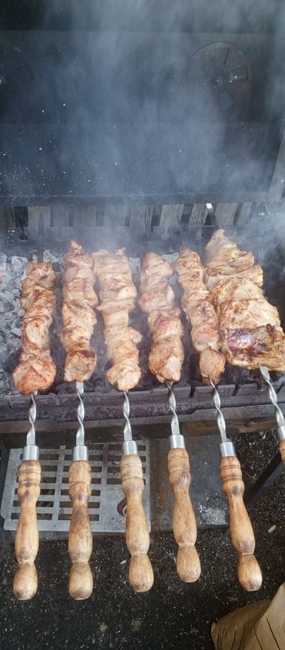 This image shows skewers of meat being grilled over hot coals. The scene is set in an outdoor grill, likely in a backyard or picnic area, given the presence of a metal grate and what appears to be dark, pebbly ground. The meat is well-browned and appears to be pork or lamb, cut into large, irregular chunks. Smoke rises from the grill, obscuring the top portion of the image, where some wooden slats are visible, possibly part of the grill's structure or a nearby fence.
The skewers are made of metal with a distinctive twisted shaft and are held by wooden handles. These handles are shaped with rounded sections and show signs of wear and use, suggesting they are part of a well-loved set of grilling tools. The meat is arranged in rows on the grill, with six skewers clearly visible in the foreground and at least two more partially visible on the right side. The coals beneath are glowing red and white, indicating intense heat.
The lighting suggests it could be daytime, with the harsh light creating shadows and highlighting the texture of the meat and wood. There are no people visible in the image, focusing the attention solely on the food and the grilling process. The overall impression is one of hearty, rustic cooking, common in outdoor gatherings. There is no visible text in the image. The location context provided, Petukhovo, Russia, hints at a possible cultural context for this type of barbecue, often referred to as "shashlik."