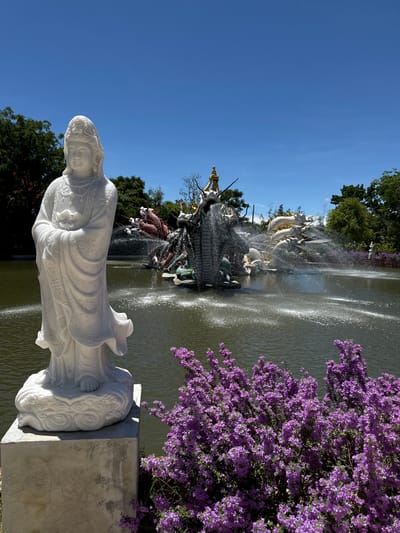 This image depicts a statue of a woman in flowing robes. The statue is white and appears to be made of marble or a similar material. It stands on a decorative base. Behind the statue is a pond or lake with a fountain. The fountain consists of multiple sculpted dragons and other figures. Water is spraying from the fountain. Purple flowers are visible to the right of the fountain. The sky is clear and blue. This is likely a tourist attraction in Bang Pu Subdistrict Municipality, Thailand.