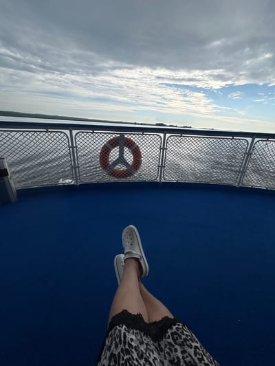 The image depicts a person's legs crossed, wearing white Crocs and a leopard-print skirt with black lace trim, resting on a blue deck. The person is on the deck of a boat, as indicated by the railing with a life preserver and a metal bin in the background. The boat is on a body of water, with a distant shoreline visible under a cloudy sky. The image does not provide information about the city of Zuevo, Russia.