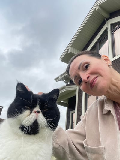 An outdoor, waist-up, eye-level, medium shot features a woman and a cat against a backdrop of a house and cloudy sky. The woman is in the right foreground, with her head tilted down towards the cat and her face turned to the right, looking at the camera with a slight smile and her eyes open. She has light skin, dark hair pulled back, and is wearing a light-colored corduroy jacket over a pink top. She has a silver circular earring in her right ear. A black and white cat is in the left foreground, with its face and chest visible. Its eyes are narrowed, and it appears to be looking down. Its fur is black and white, with black markings on its face. The cat's right ear is being gently touched by the woman's hand. The house behind them has a dark brown facade with white trim, and green-painted eaves. The sky is overcast and grey. The location is identified as the city of Solov'evka, Russia.