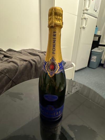 A bottle of Pommery champagne lies horizontally on a gray, textured surface. Part of a gray fabric is visible beneath the bottle. In the background, there is a white appliance and a gray storage container, suggesting an indoor environment. The image does not provide information about Katwijk, Netherlands.