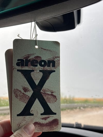 A close-up shot shows an "areon X" air freshener hanging from a car's rearview mirror. The air freshener is made of light-colored cardboard and is adorned with red lipstick kiss marks and the text "areon X" in black. The background is blurry, showing a cloudy sky and a hint of a field or landscape outside the car window. A person's fingers are visible at the bottom left, holding the air freshener. The scene is indoors, within a vehicle. The location in Komarevo, Bulgaria is not identifiable from the image.