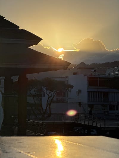 The image depicts a sunset or sunrise over the city of Papeete, French Polynesia (specified as Papeete, France by the prompt). The low sun, partially obscured by clouds, is visible in the upper center, casting a golden light and producing prominent sun rays and lens flare.

In the foreground, the dark, multi-tiered roof of a building or gazebo is silhouetted against the bright sky, occupying the upper left quadrant. The lower foreground features a reflective surface, likely wet pavement or a body of water, showing a strong, elongated reflection of the sun.

The midground reveals a cityscape. A pedestrian bridge with dark railings crosses horizontally from the lower-mid right. Below the bridge, indistinct vehicles are visible on what appears to be a road or street. Several multi-story buildings, characterized by windows, balconies, and air conditioning units, line the background of the midground. A deciduous tree with bare branches stands silhouetted against these buildings, just to the right of the foreground roof structure. One individual is visible walking on the pedestrian bridge, silhouetted against the illuminated surface.

The background consists of rolling hills or mountains under a partly cloudy sky, further away from the city structures. The overall scene is bathed in the warm, intense light of the golden hour, creating strong contrasts between illuminated areas and dark silhouettes.