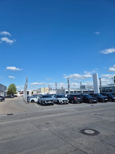 An outdoor, ground-level, vertical, wide shot captures a clear blue sky with scattered white clouds. In the lower half of the frame, a large outdoor car dealership lot is visible, filled with numerous vehicles, primarily white and black cars. The cars are parked in rows under a covered structure, suggesting a sales or display area. To the right of the cars, a tall, white sign stands, with text indicating "Audi Gebrauchtwagen" and "+plus", which translates to "Audi Used Cars". Further to the right, a building with a large red logo that reads "Audi" is visible. In the foreground, a paved asphalt surface with parking lines and a manhole cover is present. On the left side of the image, a street with a few parked cars and a tree can be seen. The overall scene depicts a car dealership on a sunny day in Landshut, Germany.