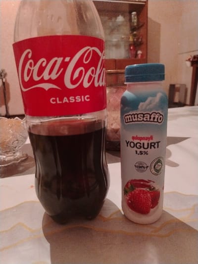 A Coca-Cola Classic bottle, partially filled, stands beside a small bottle of musaffo qulupnayli strawberry yogurt (1.5%). The bottles are positioned on a surface, possibly a table. Background elements include a glass bowl, a cabinet with items inside, and a portion of a table leg, contributing to an indoor setting.