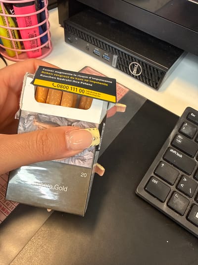 The scene depicts an indoor, office-like environment in Sint-Niklaas, Belgium, centered on a person's left hand holding an open pack of Marlboro Gold cigarettes. The hand, with a light skin tone, grasps the pack between the thumb and index finger. The fingernails are manicured with a clear or light pink gel polish base and yellow tips adorned with small, clear rhinestones. The cigarette pack is open, revealing the top portions of several brown-filtered cigarettes and a silver foil wrapper inside. The interior of the top flap features a black warning label with white text in French ("Fumer augmente le risque d'impuissance"), Dutch ("Roken vergroot de kans op impotentie"), and German ("Rauchen bedroht Ihre Potenz"), alongside the Belgian tobacco cessation helpline number '0800 111 00' and website 'tabacstop.be'.
The background consists of a dark, possibly black or dark grey, desk surface. To the upper right, a black Dell OptiPlex mini-PC is visible. A portion of a black computer keyboard, showing keys like "shift lock," "ctrl," and directional arrows, is visible on the right side of the desk, exhibiting some surface wear or dust. In the upper left, a pink mesh pen holder contains several fluorescent markers (pink, yellow, green). Partially obscured beneath the cigarette pack is a piece of paper with printed numbers, possibly a receipt or price list.