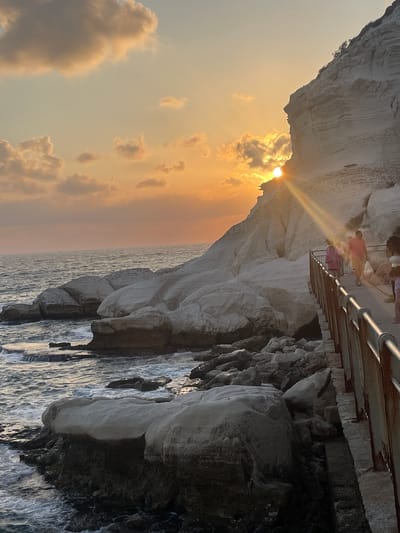 This image captures a sunset over the Mediterranean Sea at Rosh HaNikra in the Mate Asher Regional Council, Israel. The sun, partially obscured by a white cliff face, casts a warm orange glow across the sky and reflects off the water. The cliff, a prominent geological feature, is layered and appears to be made of chalk or limestone. A pathway, bordered by a weathered metal railing, runs along the cliff's edge, and several individuals, including a person in a red shirt carrying a white bag and a person in a pink jacket, are walking on it. The sea is choppy, with waves breaking against a rocky shoreline composed of large, light-colored boulders and dark, jagged rocks closer to the water. The scene is a natural coastal landscape enhanced by a man-made pathway, suggesting a tourist or recreational area.