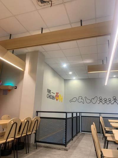 The image displays the unoccupied interior of a restaurant located in Calukan RT 1 RW 5 Keboansikep, Indonesia. The ceiling features a white acoustic tile grid with recessed circular spotlights and two linear LED light fixtures. Light-colored wooden beams are suspended decoratively from the ceiling. Walls are uniformly painted light grey. A small wooden shelf with an upward-facing teal light source is mounted on the left wall. The rear wall presents black text reading "FIRE CHICKEN" with red and yellow stylized heart and flame graphics, adjacent to a mural of black line art depicting abstract forms transitioning into stick figures of a family. The floor is covered with light-toned rectangular tiles. Seating areas include multiple light wooden tables and chairs featuring black metal frames and circular cutouts on their backs, positioned on both the left and right sides of the frame. A black metal railing with perforated circular panels partitions the left foreground seating from the mid-ground area. No individuals or active events are depicted within the scene.