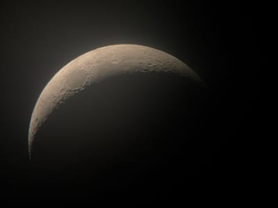 The image shows a crescent moon against a dark sky. The moon is illuminated on its right side, showing craters and surface features. The background is black, indicating nighttime. There are no people or other objects visible in the image. The scene is a close-up view of the moon, likely taken with a telescope or a camera with a powerful zoom lens. There are no visible text elements. The overall mood is serene and cosmic.