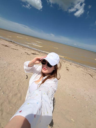 A woman is taking a selfie on a sandy beach. She is wearing a white baseball cap, sunglasses, and a white button-down shirt with colorful heart patterns over a white tank top. She is holding her hand up to shield her eyes from the sun. The background shows a murky body of water and a cloudy blue sky. The location is the beach in Port-Katon, Russia.