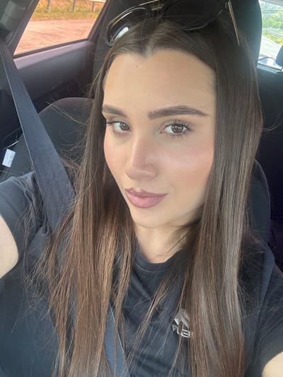 A woman with long, dark brown hair and light makeup is taking a selfie in a car. She is wearing a black t-shirt and a seatbelt. The car's interior is visible, including a black seat and a window showing an outdoor environment. The location is identified as Osikovska Lakavitsa, Bulgaria.