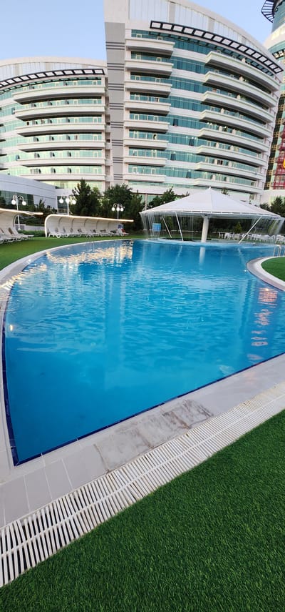 The image depicts an outdoor swimming pool area at a resort or hotel in Türkmenbaşy, Turkmenistan. A large, irregularly shaped blue swimming pool occupies the foreground and middle ground. The pool is surrounded by a light-colored tiled deck, which transitions to a white grated drainage system and then to artificial green turf on the right side of the image.

In the background, a modern, multi-story building with a curved facade and numerous windows is visible. The building appears to be made of white and glass materials. To the left of the building, several white, semi-enclosed lounge chairs with white fabric coverings are arranged on the artificial turf. White decorative lamps on poles are also present in this area.

In the middle ground, to the right of the lounge chairs, a large white canopy structure is set up over a circular area with tables and chairs. Water cascades down from the edges of this canopy, creating a decorative waterfall effect. The sky is clear and a light blue, suggesting daytime. There are no people visible in the image. The scene conveys a sense of leisure and luxury.