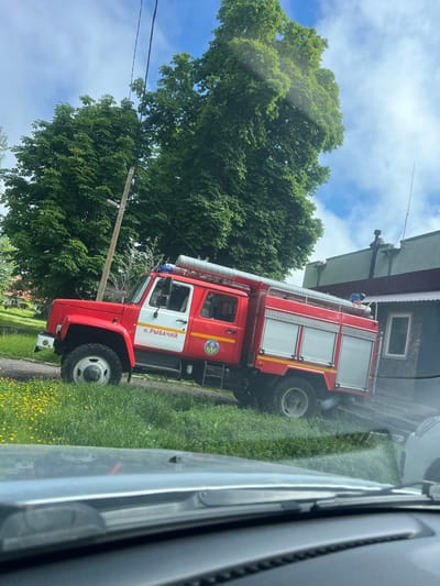 In Rybachy, Russia, a red and white fire truck is parked on a grassy area next to a dark gray building with a pitched roof. The fire truck has a large, silver hose running along its top and equipment compartments on its side. The text "п. РЫБАЧИЙ" is visible on the side of the cab. A blue flashing light is mounted on the roof of the cab. Tall green trees are in the background, and the sky is partly cloudy. The image is taken from inside a vehicle, with the dashboard and windshield wipers visible in the foreground.