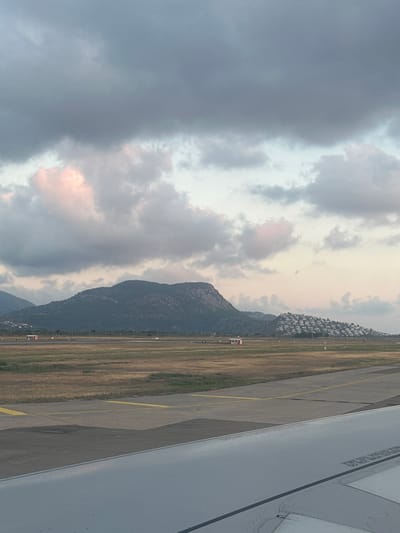 The image captures a ground-level view from an aircraft at Dalaman Airport in Dalaman, Turkey, likely at dawn or dusk. The lower right foreground shows a portion of a grey airplane wing, displaying riveted surfaces and inverted black text, including "DO NOT WALK OUTSIDE THIS AREA." The mid-ground features a concrete airport tarmac or taxiway with distinct yellow markings. Adjacent to the tarmac, a broad expanse of dry, yellowish-brown grass stretches towards the horizon. Scattered across this field are small, white and red structures, possibly airport utility buildings or navigational aids, with some ground lights visible. In the background, dark, rugged mountains dominate the landscape, covered with sparse vegetation. To the far right, on a lower hill situated in front of the main mountains, a dense cluster of white residential buildings with light-colored roofs is visible, characteristic of a hillside town or resort. The sky is largely overcast with heavy, dark grey clouds, interspersed with lighter grey and subtle pink clouds, particularly towards the lower cloud line, indicating the low sun angle. No people or active interactions are visible within the frame.