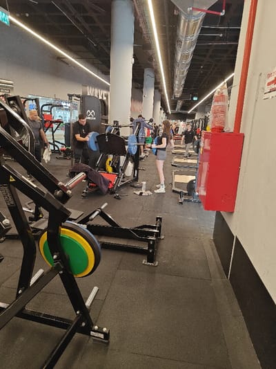 The image displays an interior view of a fitness gym in Nahariya, Israel. The facility features a dark gray rubber mat floor, light gray walls with a black kickplate, and tall, light gray concrete pillars. The ceiling is exposed with visible metallic ventilation ducts, electrical conduits, and horizontal strip lighting.

In the foreground on the left, a black Hammer Strength weight machine is partially visible, showing yellow and green weight plates. Moving to the mid-ground, a man in a black t-shirt is actively adjusting blue weight plates on a leg press machine, over which a dark purple towel is draped. To his immediate left, another man with a shaved head, wearing a dark gray t-shirt, stands holding a white towel. To the right of the man adjusting weights, a young woman with long dark hair, dressed in a dark t-shirt and gray shorts, stands holding two black weight plates. A white water bottle rests on the floor near her feet.

Further into the background, several other individuals are present and engaged in gym activities. One person in a blue hooded sweatshirt is visible near barbell racks. On the right side of the frame, a red fire safety cabinet with Hebrew text is mounted on the wall. The overall environment is well-lit and appears to be in active use by multiple patrons.