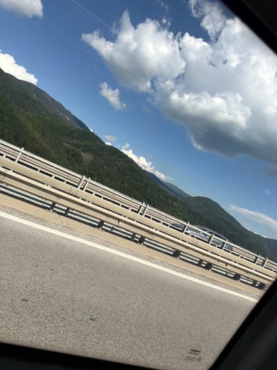 This media file captures a scenic view from a car driving on a road, likely a bridge or elevated highway, in Vrachesh, Bulgaria. The perspective is tilted, suggesting the photo was taken from a passenger's window.
The main subjects are the road itself, a metal guardrail, a densely forested mountainside, and a bright blue sky filled with fluffy white clouds. A light-colored car is visible in the distance, driving on the same road, partially obscured by the guardrail.
The scene depicts a daytime drive through a mountainous landscape. The sun is shining, casting shadows from the guardrail onto the road, indicating a clear and pleasant day. The lush green of the trees suggests it is likely spring, summer, or early autumn. The mountains are tall and imposing, with the road winding along their side.
Notable details include the modern construction of the guardrail, composed of parallel metal bars and concrete supports. The asphalt road has a clear white line marking the lane. The clouds are dynamic, with some appearing wispy and others more substantial, hinting at a light breeze. There are no visible signs of text in the image. The overall mood is one of peaceful travel and appreciation for natural beauty.