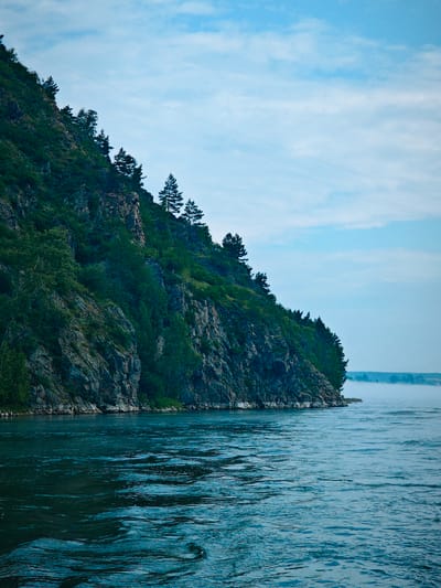This image captures a wide view of the Yenisei River in Atamanovo, Russia, with a steep, tree-covered rocky cliff face dominating the left side of the frame. The river occupies the lower half of the image, its surface appearing dark blue and rippled, suggesting a moderate current. Mist or fog hovers near the distant riverbank on the right, partially obscuring the horizon and a faint outline of trees. The sky above is a pale blue, with scattered wispy clouds. Several thin, dark lines, likely power lines, stretch diagonally across the upper right portion of the sky and over the cliff. The rocky cliff is textured with various shades of gray and brown, interspersed with dense green foliage and scattered evergreen trees. The scene is devoid of human presence and any visible structures, presenting a natural, somewhat rugged landscape.