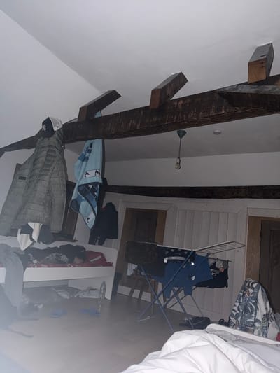 This indoor scene depicts an attic room with exposed dark wooden beams on a white ceiling. A clothes-drying rack holds blue clothing, and a table with various items, including clothing and a water bottle, sits nearby. Clothing items are scattered around the room. This image was likely taken in Lahnau, Germany.