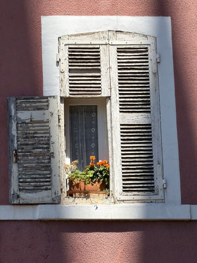 The image shows a close-up of an old, weathered window on a pink stucco building. The window has white wooden shutters, one of which is partially open, revealing a lace curtain behind it. A planter box sits on the windowsill, filled with green foliage and small orange flowers. The building is located in the city of Voiron, France. The lighting suggests it is daytime, with sunlight casting shadows on the wall and window.
