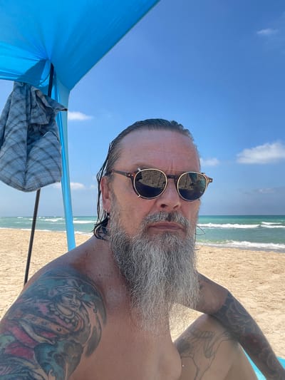 A man with long gray hair pulled back, a long gray beard, and sunglasses is seated on a sandy beach in Ga'ash, Israel, under a blue beach canopy. He is shirtless, revealing multiple colorful tattoos on his arms and a sketch-like tattoo on his leg. The ocean with moderate waves is visible in the background, under a clear blue sky with scattered clouds. A grey and blue plaid shirt is draped over a pole of the canopy. The man is looking directly at the camera.