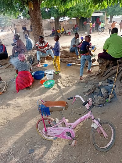 This image depicts an outdoor communal area in Nata-Ala, Nigeria. The scene is characterized by dirt ground, strong daylight casting shadows, and a large tree with prominent exposed roots dominating the central foreground.
In the foreground, a pink child's bicycle, branded "VICJOY" and "GEONIKS," rests on the ground. To the left, a woman wearing a grey top and a red wrap is seated on a low stool, tending to a small, smoldering fire contained by rocks. Adjacent to her are a blue plastic basin, a white plastic bucket, an orange bowl, and a green woven basket.
Multiple individuals are seated on the tree roots. From left to right: an older man in a brown robe and cap; another older man in a light blue robe and cap; a man in a red, white, and black striped polo shirt and light blue jeans, interacting with a young boy in a blue shirt and yellow track pants; a man in a grey t-shirt holding a young child in a blue top and jeans on his lap, both holding cups; and a man in a lime green t-shirt seated with his back partially to the camera.
In the mid-ground and background, additional people are present near single-story structures, some with thatched roofs and others with corrugated metal roofs. One individual is visible riding a bicycle past these buildings. The overall scene portrays a casual gathering with various daily activities underway.