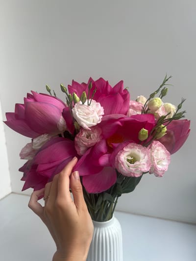 A hand is holding a bouquet of pink lotus flowers and white eustoma flowers in front of a white background. The stems are tied together and appear to be in a white vase. The hand has a ring on the finger. There is no indication of the location of Nam Natural Heritage Area Trang, Vietnam.