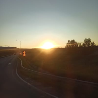 The image captures a sunset scene on a highway with gentle curves, likely located near Nicolae Bălcescu, Romania. The sun is low on the horizon, casting a warm, orange glow and creating lens flare effects. Two cars are visible on the road, one in the distance and another closer to the foreground. Traffic signs, including speed limit signs, are present on the side of the road. The landscape is characterized by rolling hills covered in vegetation, with a few trees silhouetted against the sky. The sky transitions from a pale blue at the top to a soft yellow near the horizon. The overall atmosphere is serene, with the natural beauty of the sunset dominating the scene.