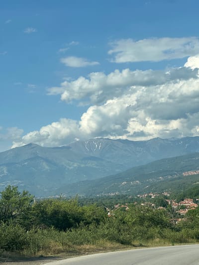The image presents a panoramic daytime view of a mountainous landscape, identifying the location in the vicinity of Barakovo, Bulgaria. The upper portion of the frame is dominated by a bright blue sky interspersed with scattered white and light gray cumulus clouds. A prominent range of large mountains, partially snow-capped or featuring light rock on their peaks, extends across the background. Successive layers of mountains and hills covered in dense green foliage recede into the distance.
In the midground, nestled among the forested slopes, a cluster of buildings with distinctive reddish-orange tiled roofs marks a village, specifically identified as Barakovo, Bulgaria. This settlement is surrounded by an abundance of green trees and vegetation. The foreground consists of dense, unmanicured green shrubs, bushes, and several deciduous trees lining the edge of a paved asphalt road. The road curves into the lower right corner of the frame, with its surface visible. The scene is illuminated by natural daylight, indicating clear conditions despite the cloud presence. No individuals or animals are visible.