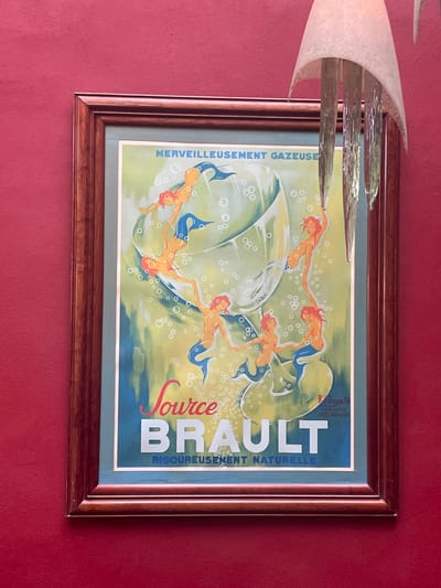 A framed vintage advertisement hangs on a red wall. The advert features mermaids swimming inside a glass of bubbly water. The text "MERVEILLEUSEMENT GAZEUSE" is on the left, while "Source BRAULT RIGOUREUSEMENT NATURELLE" is to the right. A modern, sculptural light fixture partially obscures the left side of the frame. The frame itself is brown wood with a dual-toned design.