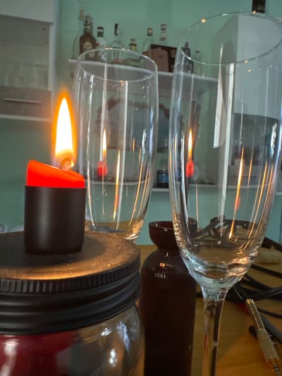 This media file shows two clear glass flutes. One flute is placed upright, and the other is inverted over it. A flame from a lighter is positioned beneath the inverted flute, creating a warm glow. The lighter has a black body and a bright orange tip. The scene appears to be indoors, possibly a kitchen or dining area, given the presence of shelves with bottles in the background. The lighting suggests it's not daytime. The overall impression is one of a close-up shot focused on the interaction of the flame and the glassware.