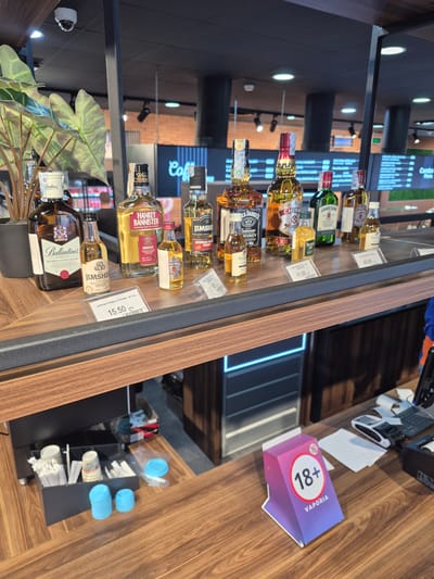 The image shows a display of alcoholic beverages on a wooden shelf, likely inside a store in Berbuki, Georgia. The focus is on various bottles of whiskey, including brands like "Ballantine's", "Jim Sher", "Hankey Bannister", "Jack Daniel's" and "Chivas Regal". Price tags are visible next to some of the bottles. A small indoor plant sits at the front of the display. In the background, there is a cash register and a sign with "18+" and "Vaporia" branding. A stack of disposable cups is also visible. The overall environment suggests a retail setting, possibly a supermarket or liquor store.