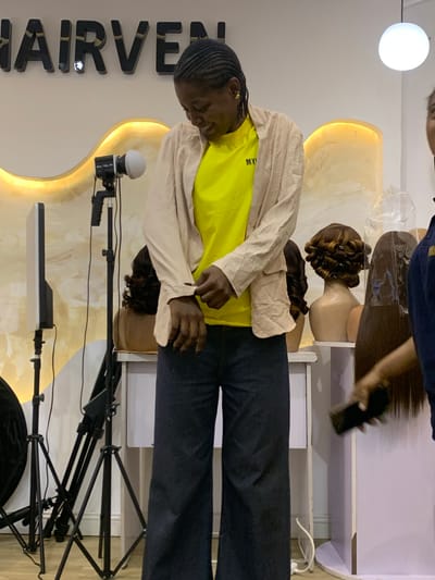 The image captures an indoor scene, likely a hair salon, beauty studio, or retail space in Animashawun, Nigeria. The central subject is a person, appearing to be a young individual, standing and looking downwards. They have dark skin and braided hair, wearing a bright yellow t-shirt with "MY VE" partially visible text, and a light beige or cream-colored long-sleeved shirt or jacket over it, paired with dark trousers.
In the background, there are modern, curved display units or shelves, illuminated from behind with a warm yellow light. Several mannequin heads are visible on these displays, showcasing various wigs or hair extensions. One wig is styled with prominent curls, while another is covered in clear plastic wrapping.
To the left of the central figure, prominent large, dark letters spell out "HAIRVE" against a white wall, suggesting the name of the establishment or brand. In the foreground, photography or videography equipment is set up, including a black tripod stand holding a long, rectangular LED panel light and another smaller, spherical light source.
Towards the top right of the image, a hand belonging to another partially visible person is seen holding a mobile phone, possibly filming or taking a picture. A bright white circular light fixture hangs from the ceiling. The overall setting appears to be designed for showcasing hair products or services, possibly during a promotional event or photoshoot.