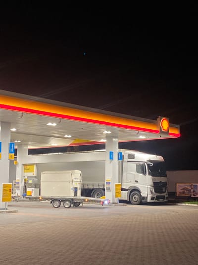 At night, a white semi-truck is parked at a Shell gas station in Horbów-Kolonia, Poland. The truck is positioned under the gas station canopy, which is illuminated with red and yellow lights. In front of the truck, a flatbed trailer is loaded with a large, rectangular, white container. The trailer has two axles and four wheels. The gas station appears to be equipped with multiple fuel pumps and signage, including a yellow sign indicating "Shell Diesel". The ground is paved with cobblestones. In the background, a billboard displays an image of a person. The sky is dark and appears clear.