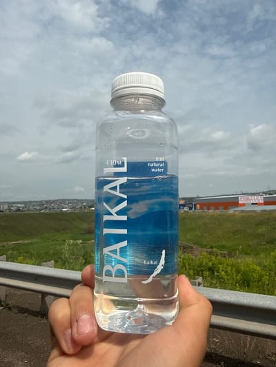 A person's hand is holding a clear plastic bottle of Baikal water. The bottle has a white cap and a blue label with white text. The label reads "BAIKAL", "430M", and "Still natural water". A silhouette of Lake Baikal is also visible on the label. The bottle is in focus, and the background is blurred. The background shows a cloudy sky, green grass, some buildings, and a highway guardrail. The scene appears to be outdoors, possibly in a suburban or rural area near the city of Solontsy, Russia, given the presence of industrial buildings and the clear sky. The image is taken from a low angle, looking up at the bottle and the sky.
