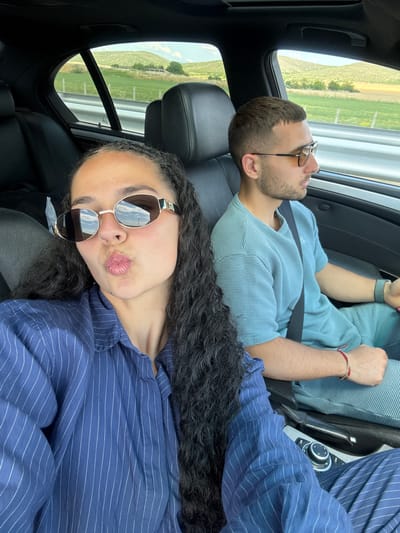 A medium shot from the interior of a car shows two people. In the foreground, a woman with long, dark curly hair and wearing a blue pinstripe shirt and oval sunglasses puckers her lips towards the camera. To her right, a man with short brown hair, a beard, and sunglasses wears a textured blue t-shirt. He is looking to his right. The car's interior includes black leather seats and a center console with a dial. Through the car window, a rural landscape with green hills and a cloudy sky is visible. The location is Polsko Padarevo, Bulgaria.