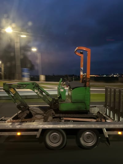 The scene depicts a green mini-excavator secured on a silver dual-axle flatbed trailer, in motion on a multi-lane roadway in Hoofddorp, Netherlands, during twilight or nighttime hours. The excavator features a black seat, control levers, and an orange rollover protection structure (ROPS) extending upwards. The excavator's arm, bearing the white "bam" logo, is folded and rests over the front section of the trailer. Visible tracks of the excavator are on the trailer bed. Stacked metal grates or crates are secured to the right rear portion of the trailer. The trailer displays illuminated amber side marker lights, and its wheels exhibit rotational blur. The background, largely blurred due to motion, shows multiple overhead streetlights with bright glare, dark supporting structures, a blurred blue rectangular sign, and distant urban or industrial lights on the horizon under a dark blue, cloudy sky. No individuals are visible.