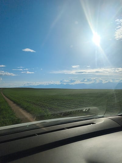 A sun-drenched view from inside a vehicle captures a vast, green agricultural field stretching towards distant, hazy mountains under a bright blue sky. A dirt road curves into the field from the left, disappearing into the horizon. The sun, high and brilliant, casts lens flares and washes out a portion of the sky on the right. Wispy white clouds are scattered across the sky, with a larger cluster of clouds above the mountain range. The foreground of the image is dominated by the interior of a vehicle, with a dark, textured dashboard and windshield reflecting the landscape. The location is identified as Kharamodun, Russia.