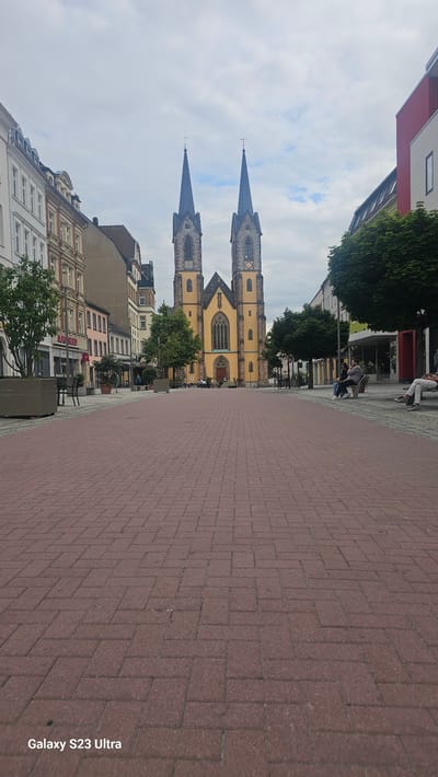 The image depicts a view of the Marienkirche in Hof, Germany, from a cobblestone plaza. The church, with its prominent yellow facade and twin spires, is centrally located in the background. The plaza in the foreground is paved with red bricks arranged in a herringbone pattern. Several people are visible in the scene: two individuals are seated on a bench on the right side of the plaza, and a few more are seated at tables outside a building on the left. Trees are planted along the sides of the plaza and in planters, adding greenery to the urban environment. The sky is overcast with scattered clouds. The buildings lining the plaza exhibit varied architectural styles. The bottom left corner of the image contains a watermark reading "Galaxy S23 Ultra".
