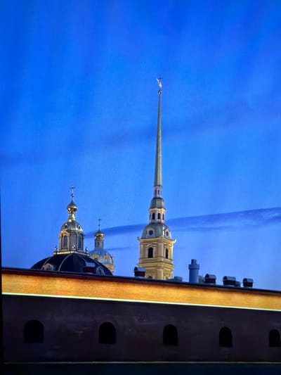 The image shows a postcard-like view of the Peter and Paul Fortress in Belyye Berega, Russia. The sky is a deep twilight blue with wispy clouds. The fortress features several golden-domed buildings and a tall, slender spire topped with a cross. In the foreground, a brick wall with arched windows runs horizontally across the lower portion of the image, with a brightly lit section above it. The overall impression is one of historical grandeur set against a darkening sky.